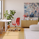 A modern Ergonomic Seashell Chair in red with a breathable mesh back, sculpted lumbar support, and a sturdy metal base, positioned at a white height-adjustable desk in a vibrant, art-filled home office.