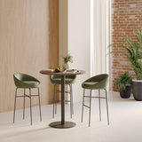 Round walnut bistro bar table with green upholstered counter stools beside brick wall and indoor plants