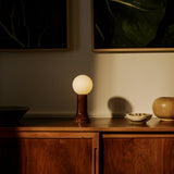 Shore table lamp with mouth-blown amber recycled glass base and glowing matte bulb, styled on a walnut sideboard in a softly lit modern interior.