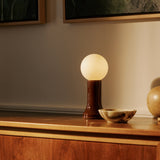 Shore table lamp with mouth-blown recycled glass base in deep blue, glowing matte bulb, styled on a wooden cabinet against a warm-toned wall.