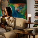 Shore table lamp with amber recycled glass base and glowing bulb on a wooden side table, beside a woman reading on a beige sofa in a cozy living room.