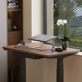 Slim silver laptop stand on wooden desk by window with orchid plant, glass of water, and modern home office decor