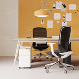 Urbanica six person office workstation desk styled in a warm workspace with mustard accent wall, black mesh chairs, and a white mobile pedestal drawer beneath the table