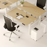 Close-up of the Urbanica Two-Person Standing Office Desk featuring sleek height-adjustable wood surfaces, ergonomic mesh chairs, and under-desk mobile storage cabinets