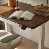 Close-up of the Urbanica Mini Standing Ergonomic Office Desk featuring a walnut wood top, white frame, and open notebook in a cozy workspace setting
